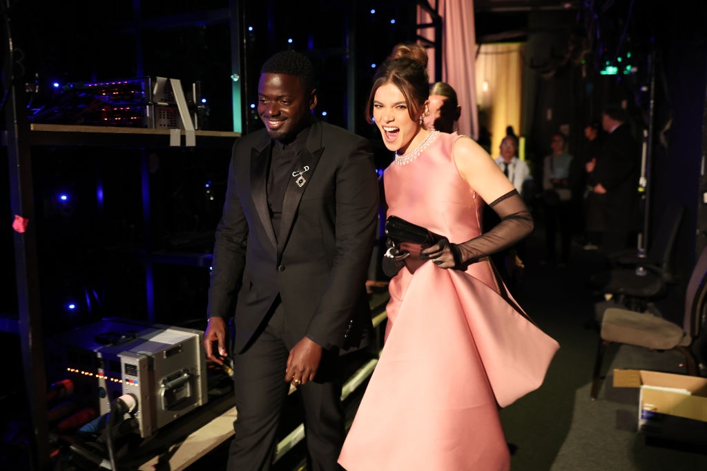 Man in a classic tuxedo and woman in an elegant sleeveless gown with gloves, smiling backstage at an event