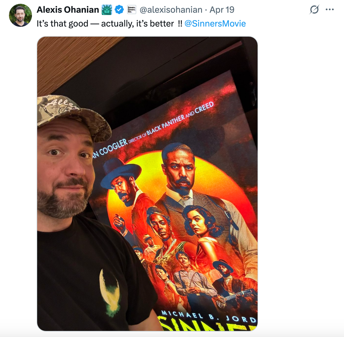 Man in a t-shirt and cap poses beside a &quot;Sinner&quot; movie poster featuring actors in dramatic expressions