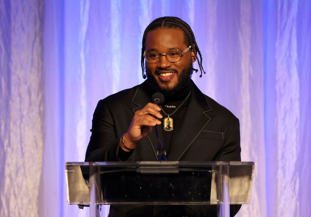 A person with braided hair, wearing a black suit and necklace, speaks at a podium on stage