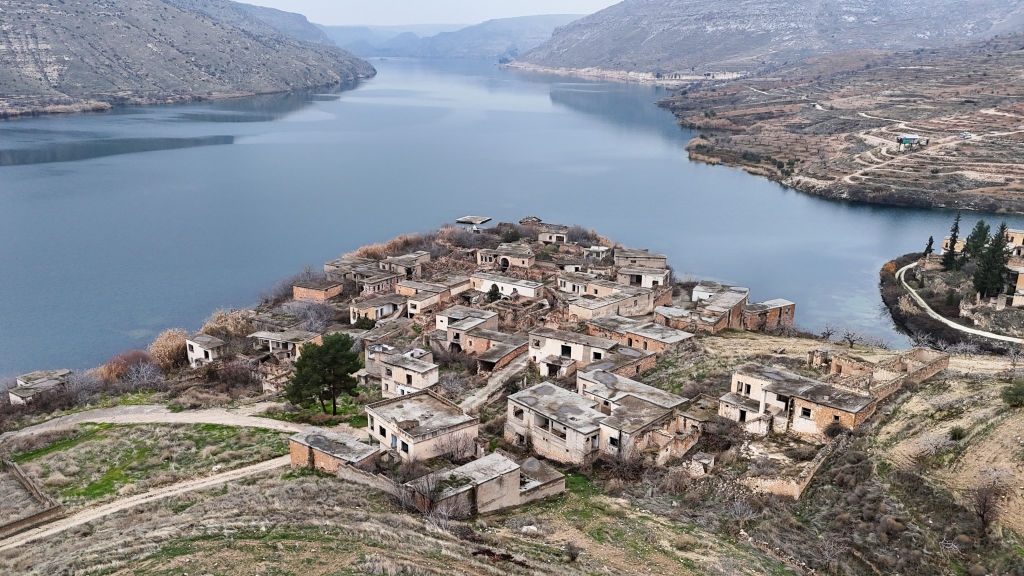 Aerial view of an abandoned village with decaying buildings by a wide river, surrounded by hills and sparse vegetation