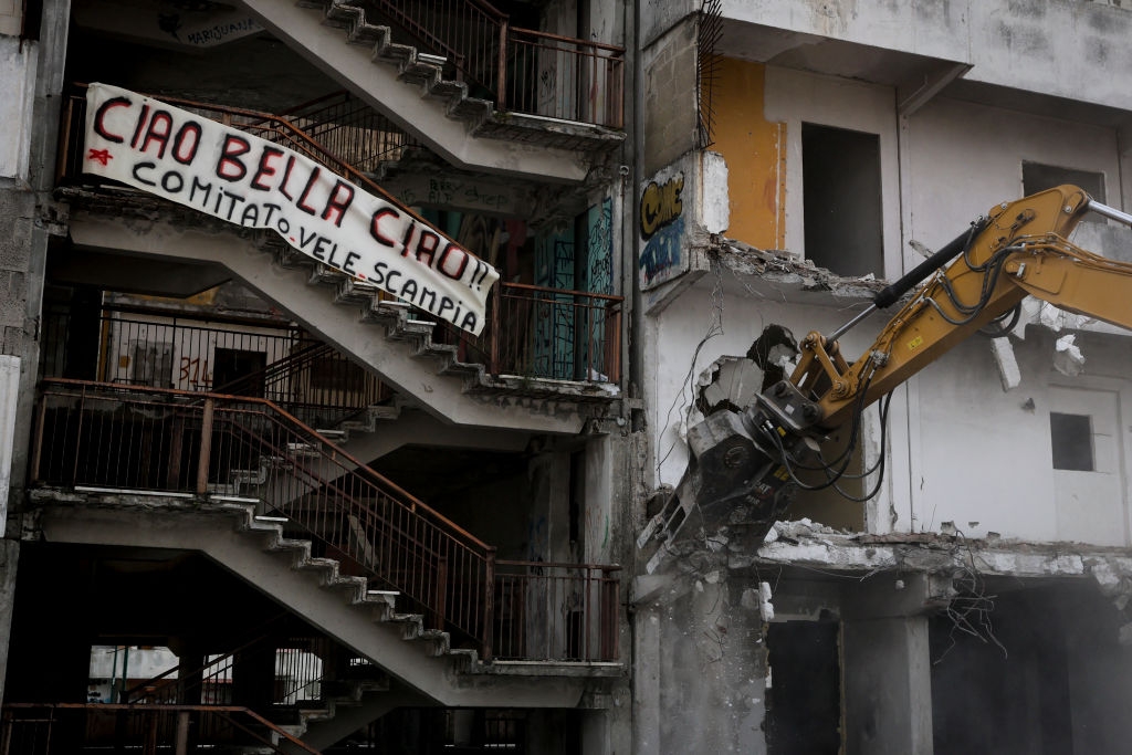 A demolition excavator tears down part of a worn building with a banner that reads "Ciao Bella Ciao, Comitato Vele Scampia"