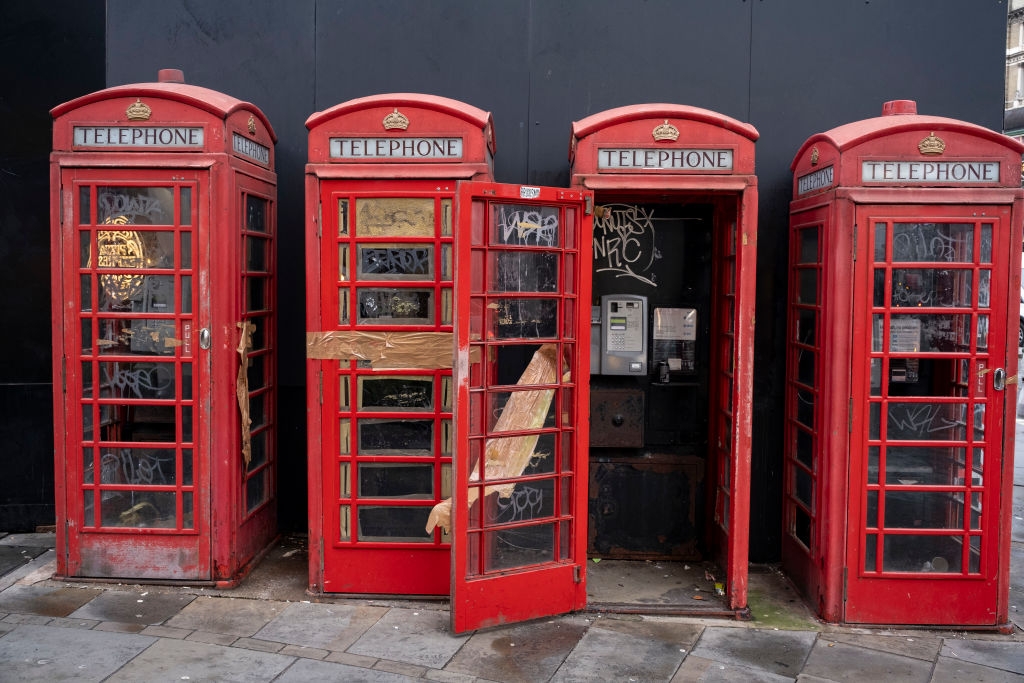 Four traditional British phone booths, one with an open door, show signs of wear and graffiti, standing on a city sidewalk. They're taped up