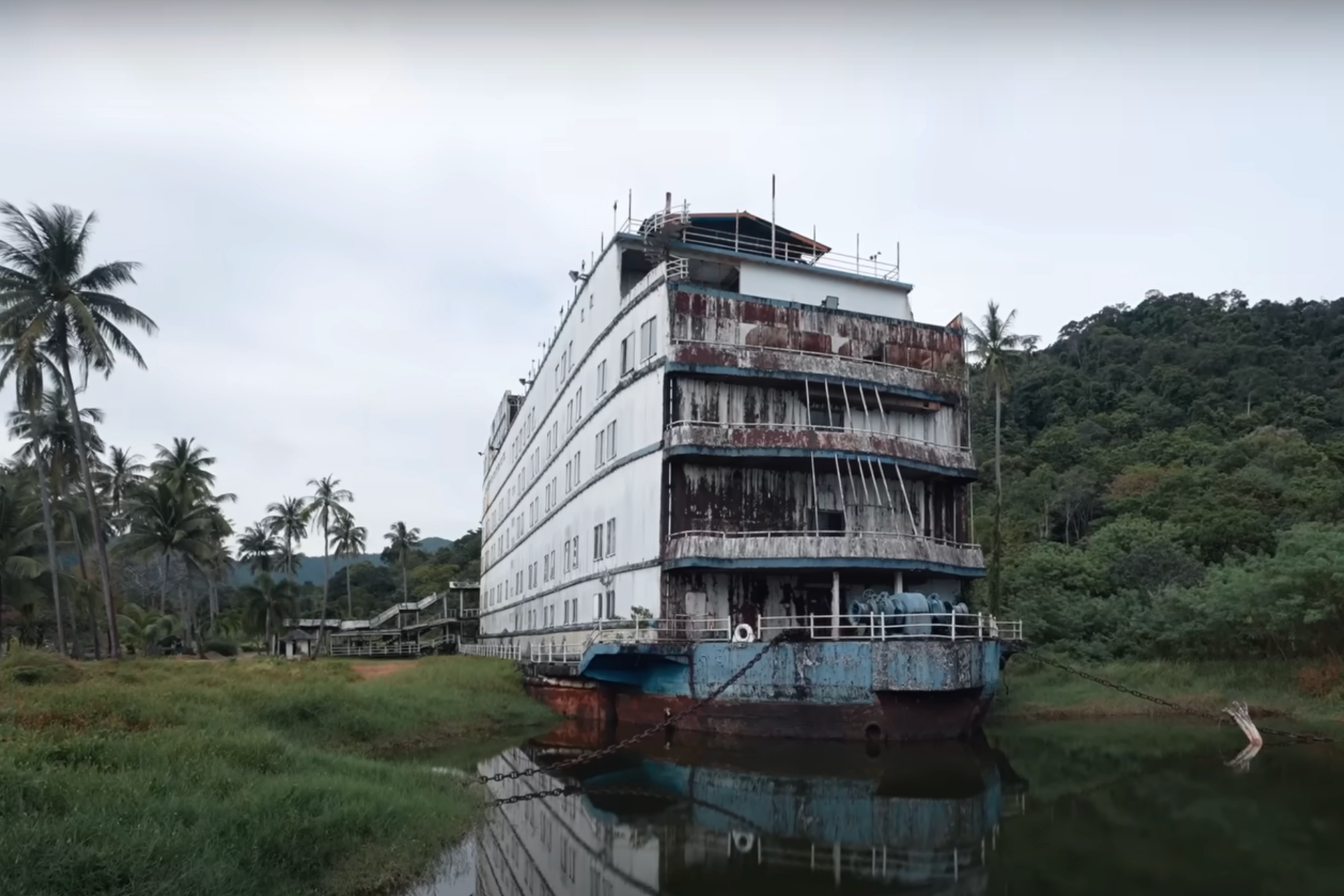 Large abandoned ship on land surrounded by trees with rust and wear, reflecting in a small body of water
