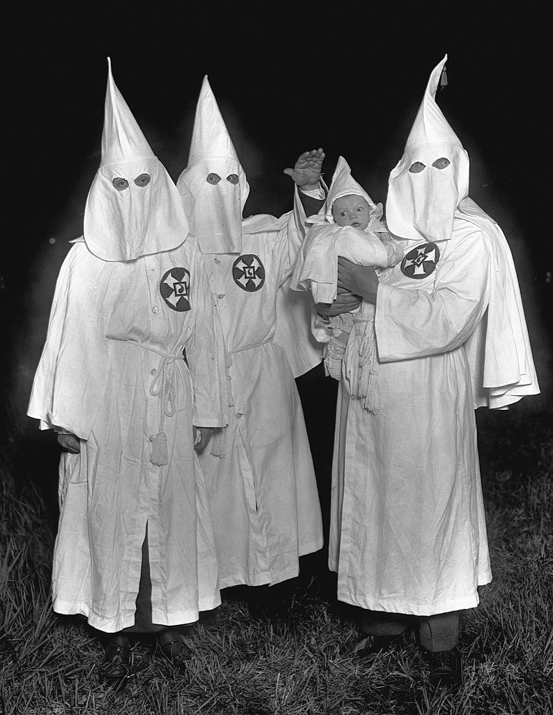 Four individuals in white hooded robes with pointed hats and a cross emblem standing in a grassy area, one holding a baby also in similar attire