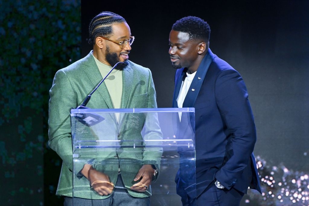 Two men speaking at a podium during an event. One wears a blazer and turtleneck; the other wears a suit and shirt, smiling at each other