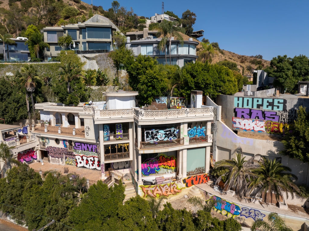 A large, abandoned house covered in colorful graffiti, surrounded by lush greenery and modern homes nearby