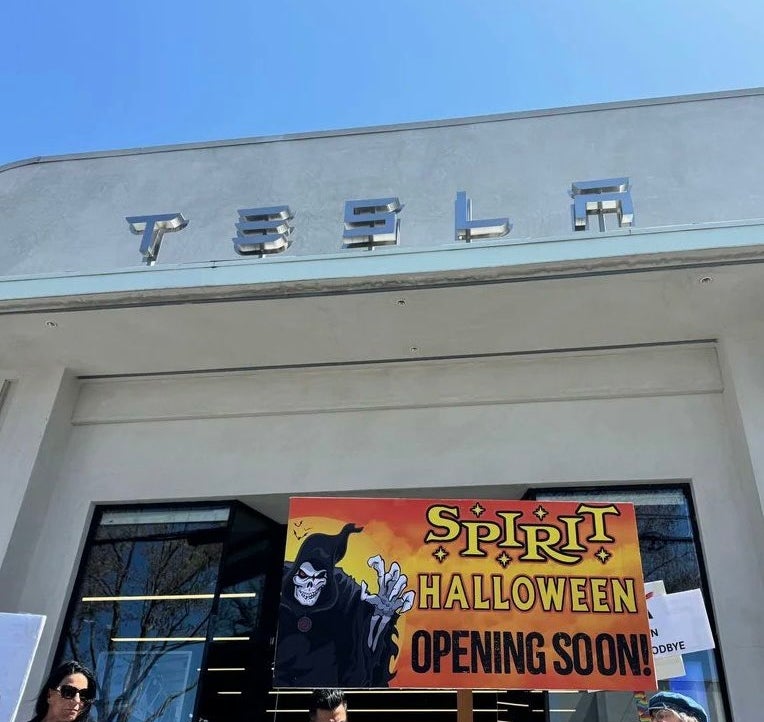 People hold a &quot;Spirit Halloween Opening Soon&quot; sign outside a Tesla store