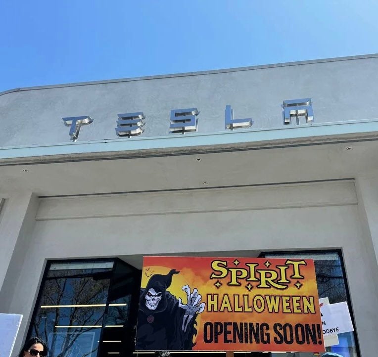 People hold a "Spirit Halloween Opening Soon" sign outside a Tesla store