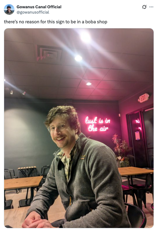 Person sitting in a café with a neon sign in the background reading "lust is in the air."