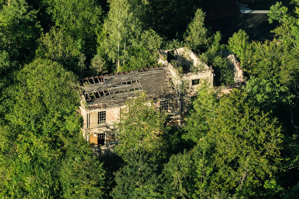 Aerial view of an abandoned stone building with wooden roof falling into disrepair surrounded by dense forest