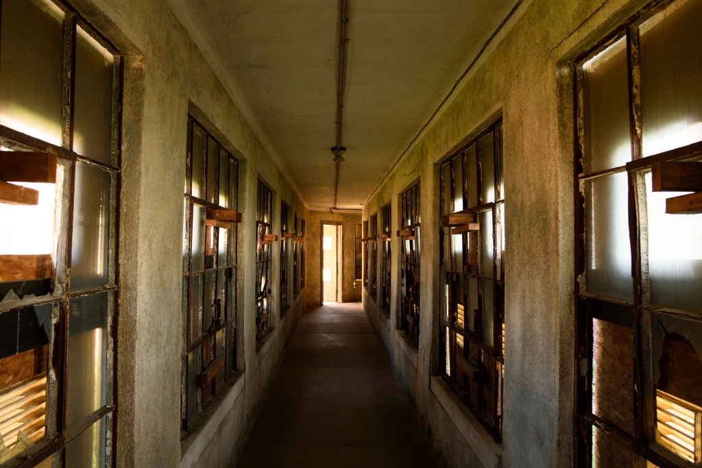 Narrow corridor with broken windows and peeling walls, leading to a door with light filtering through, creating an eerie, abandoned atmosphere