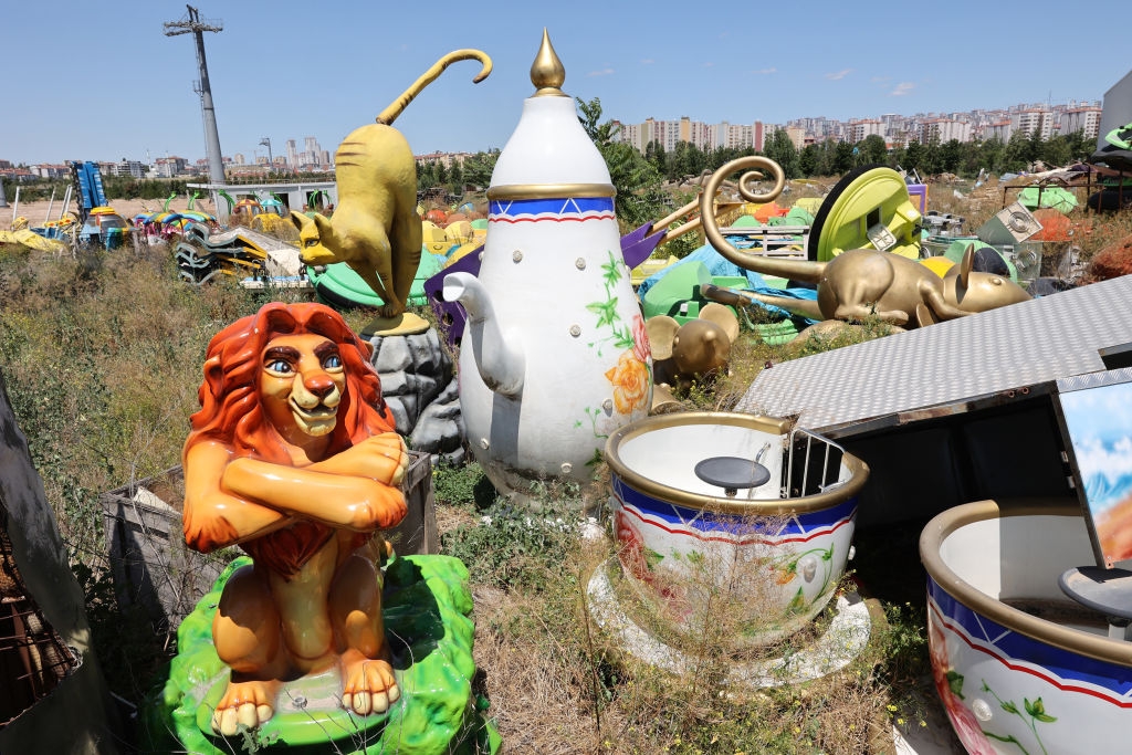 Overgrown field with with large character and teapot figures, including a lion statue sitting cross-armed, among various colorful amusement park props