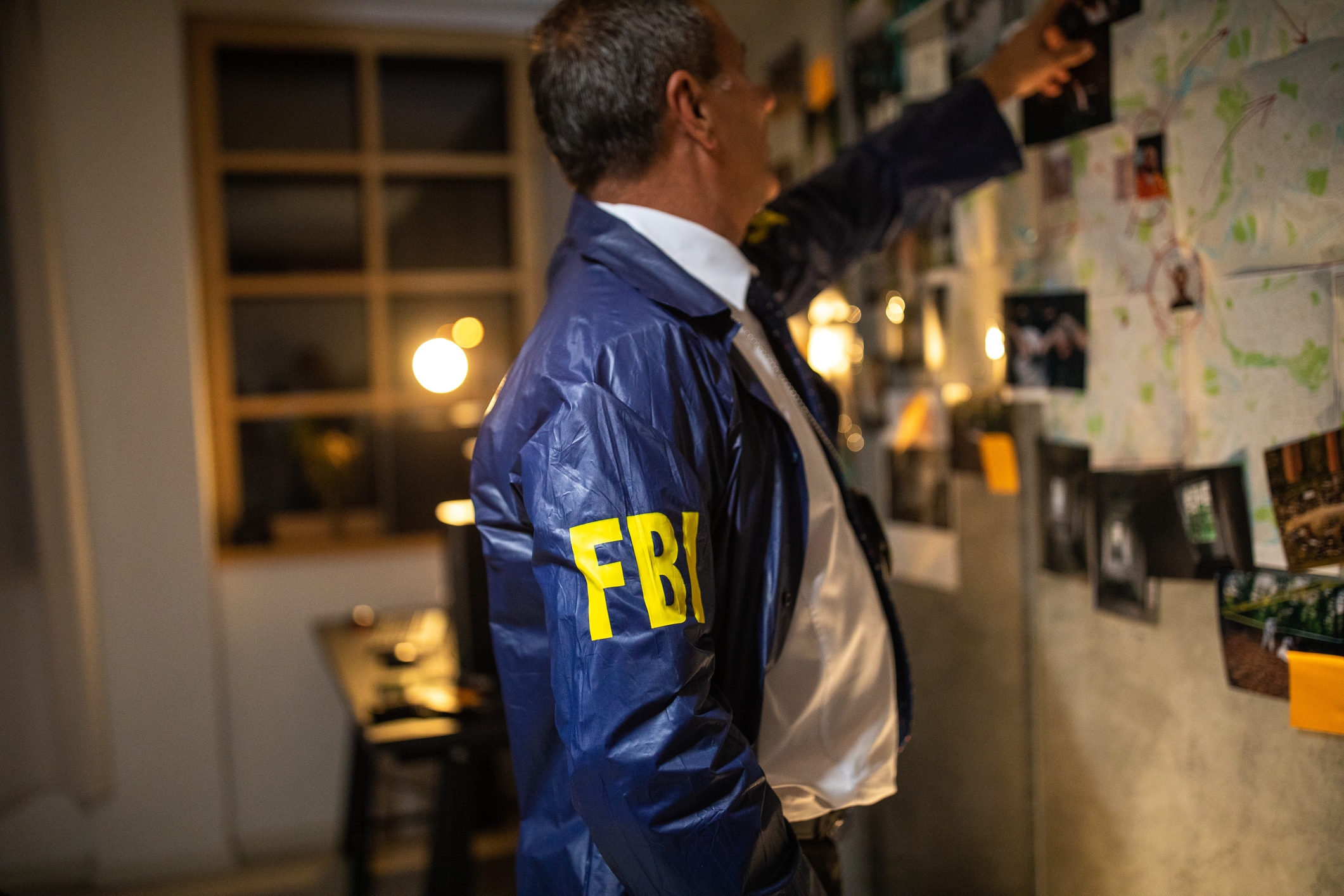 A person in an FBI jacket examines a board covered with photos and documents in a dimly lit room