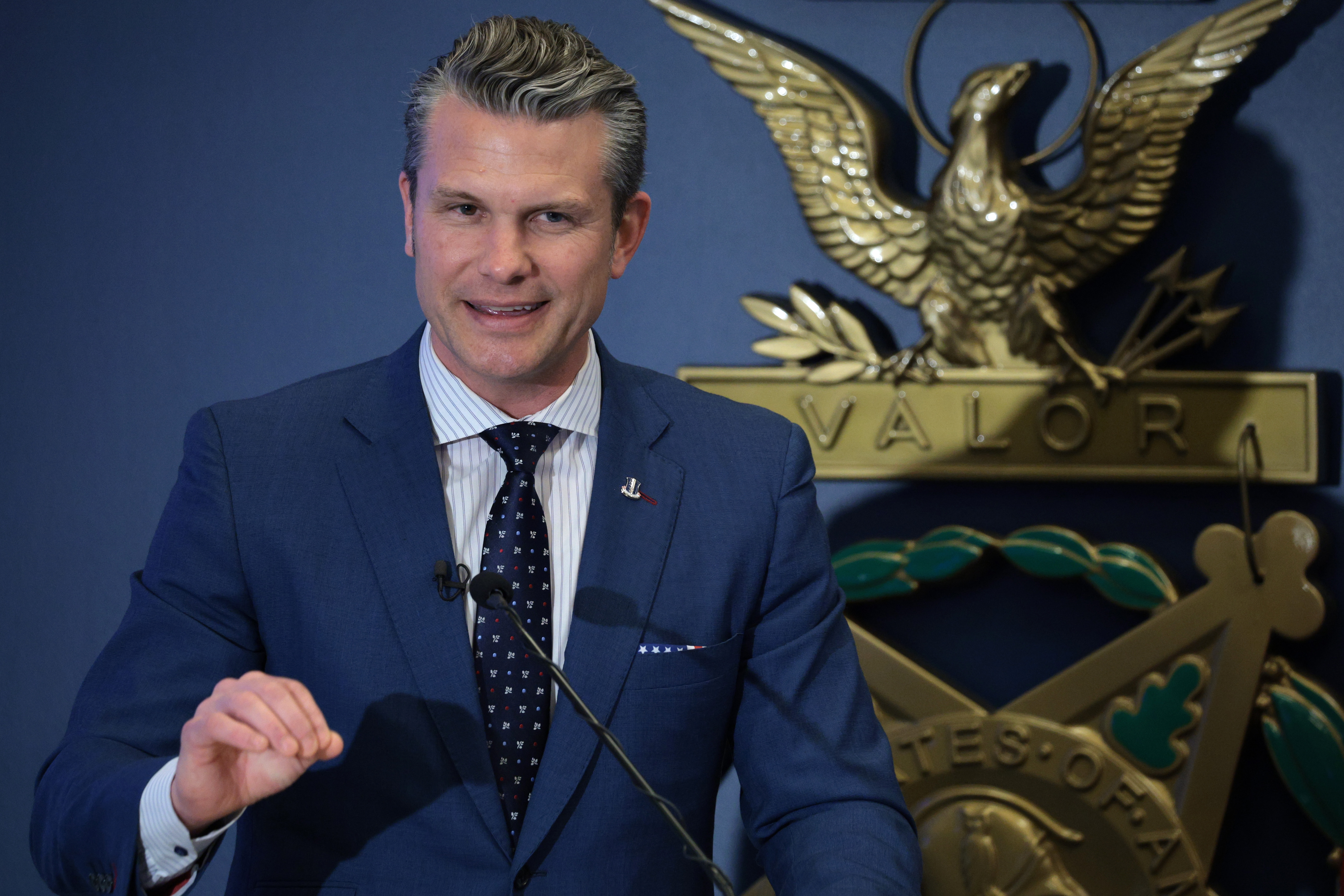 Man in a suit gives a speech at a podium with a decorative emblem featuring an eagle and the word "VALOR" behind him