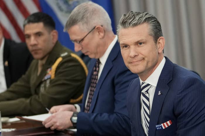 A man in a suit and striped tie sits at a conference table with others in military and formal attire, engaged in a meeting