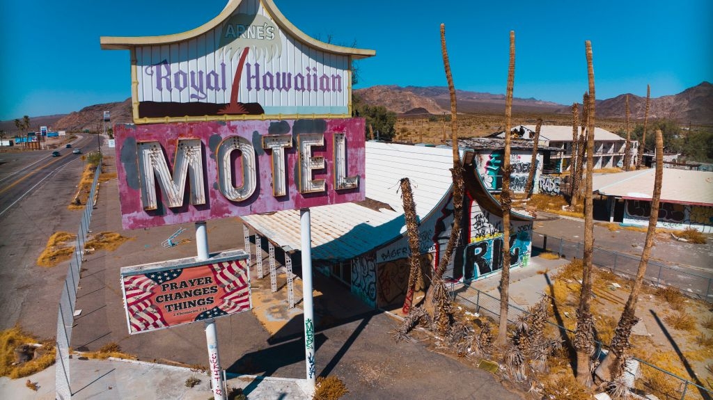 Abandoned motel with a large, worn sign reading "Royal Hawaiian Motel" and a smaller American flag banner sign that says "prayer changes things." The area shows signs of decay and disuse, with graffiti on buildings