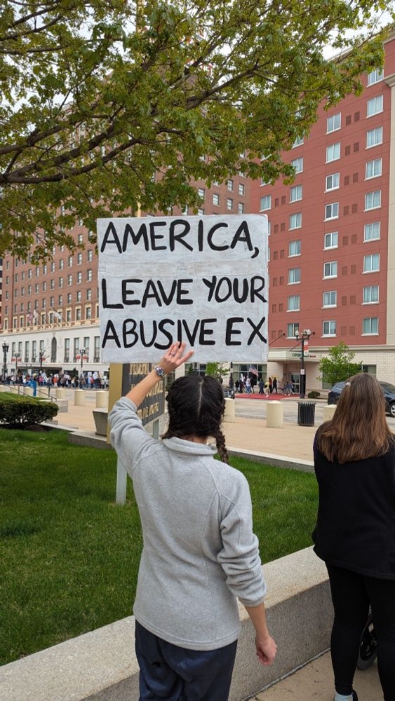 Person holding a protest sign reading, "America, leave your abusive ex," in an urban setting