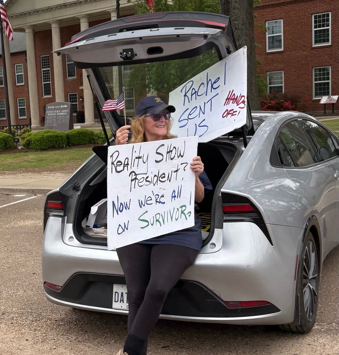 A woman sits in a car trunk with signs reading &quot;Reality Show President? Now we're all on Survivor&quot;