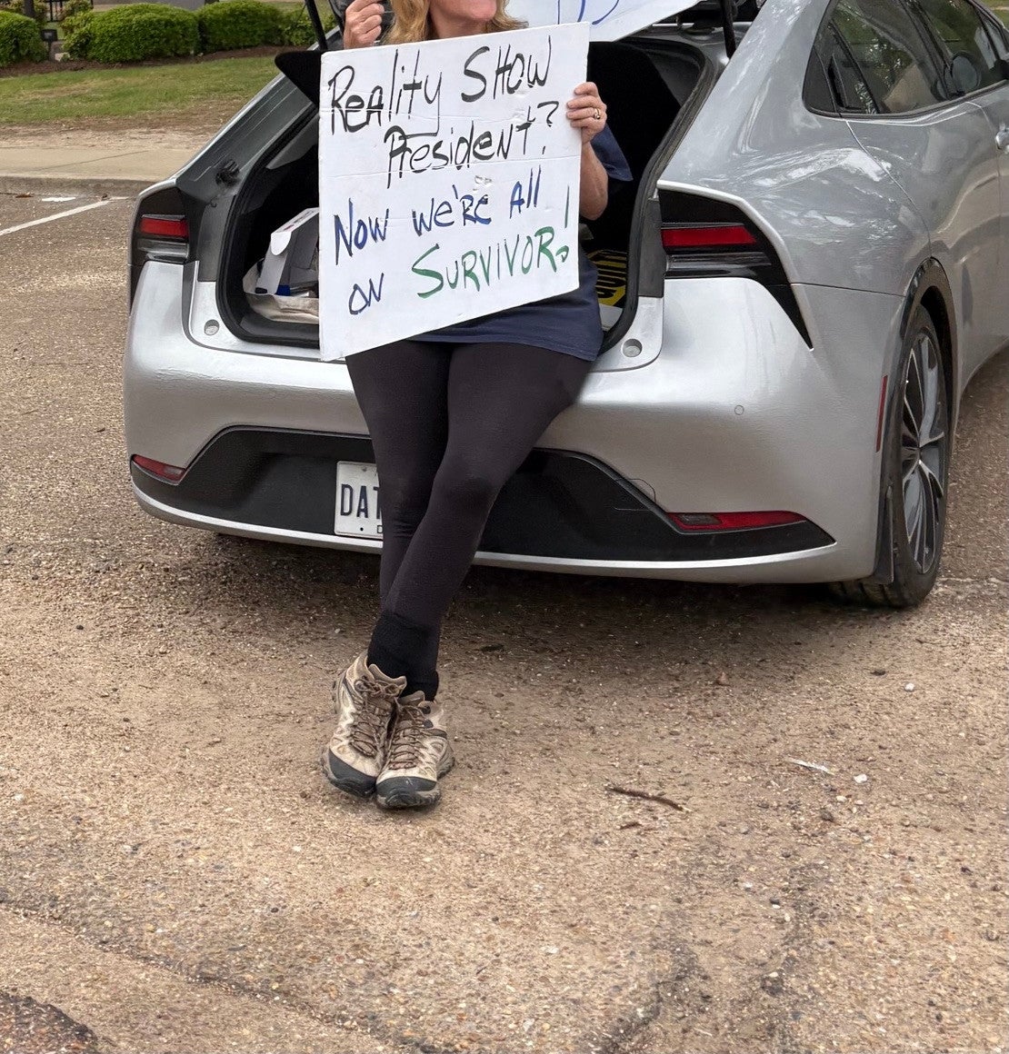 A woman sits in a car trunk with signs reading "Reality Show President? Now we're all on Survivor"