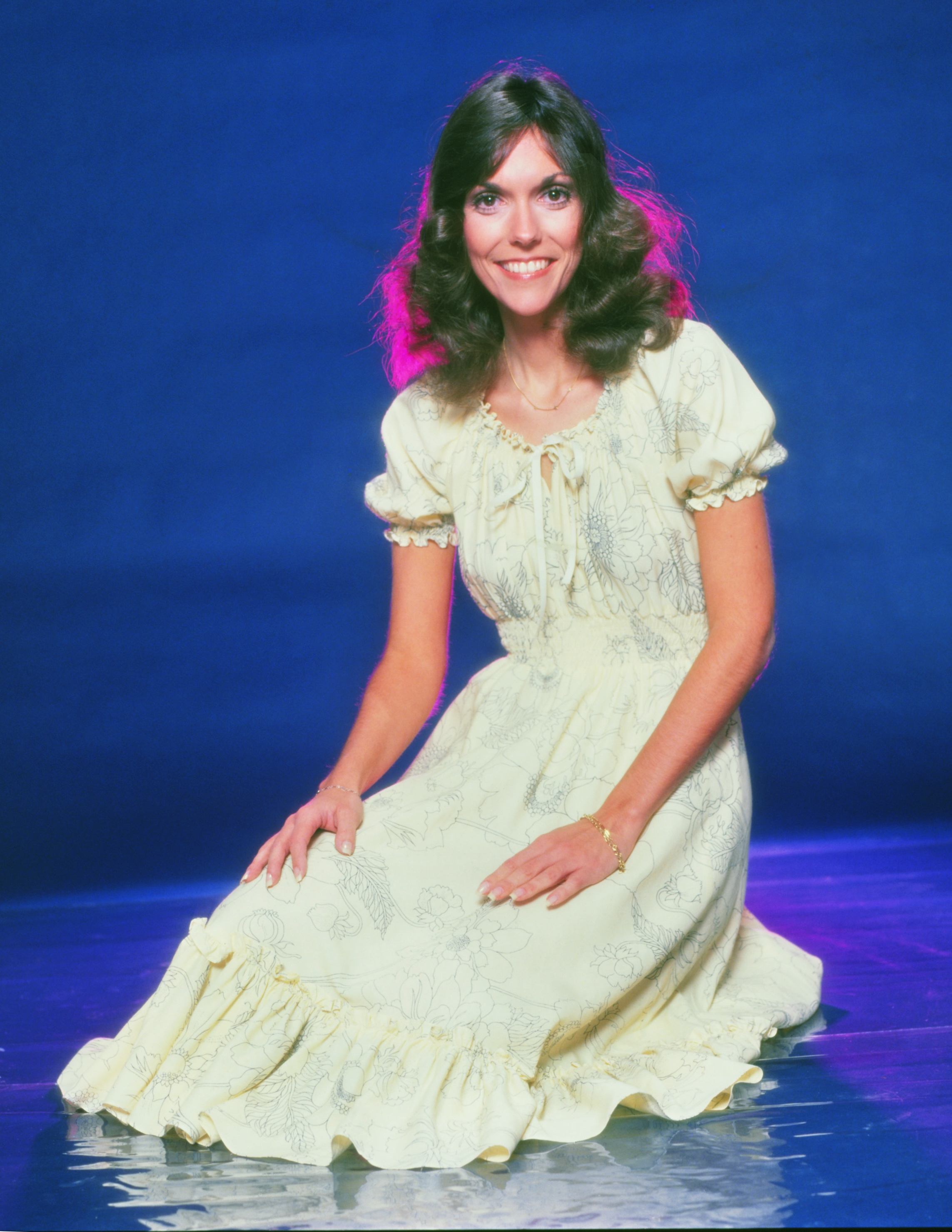 Person in a vintage floral dress kneeling with hands on lap, smiling at the camera