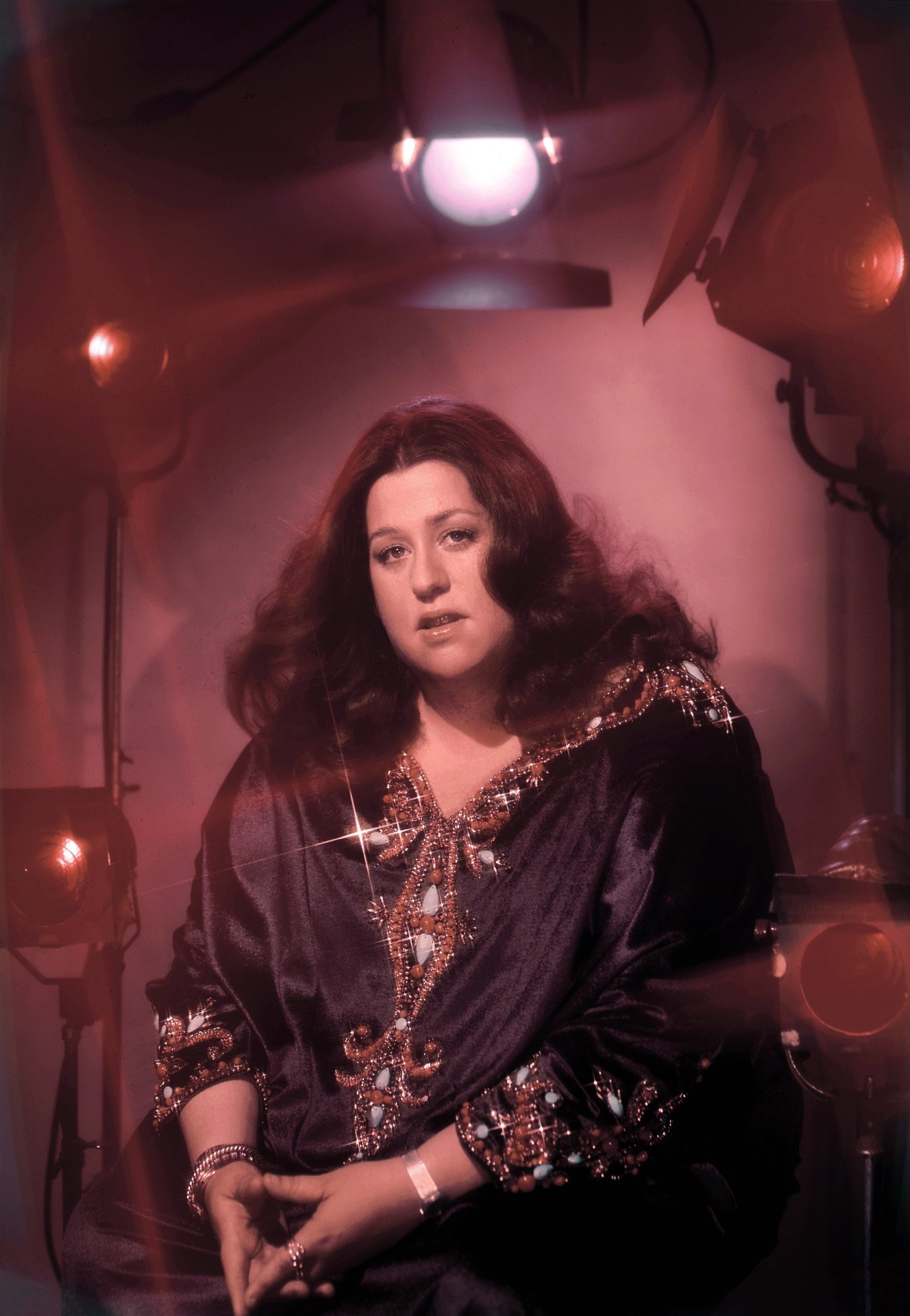Person in a studio wearing an embroidered tunic, seated amid stage lights