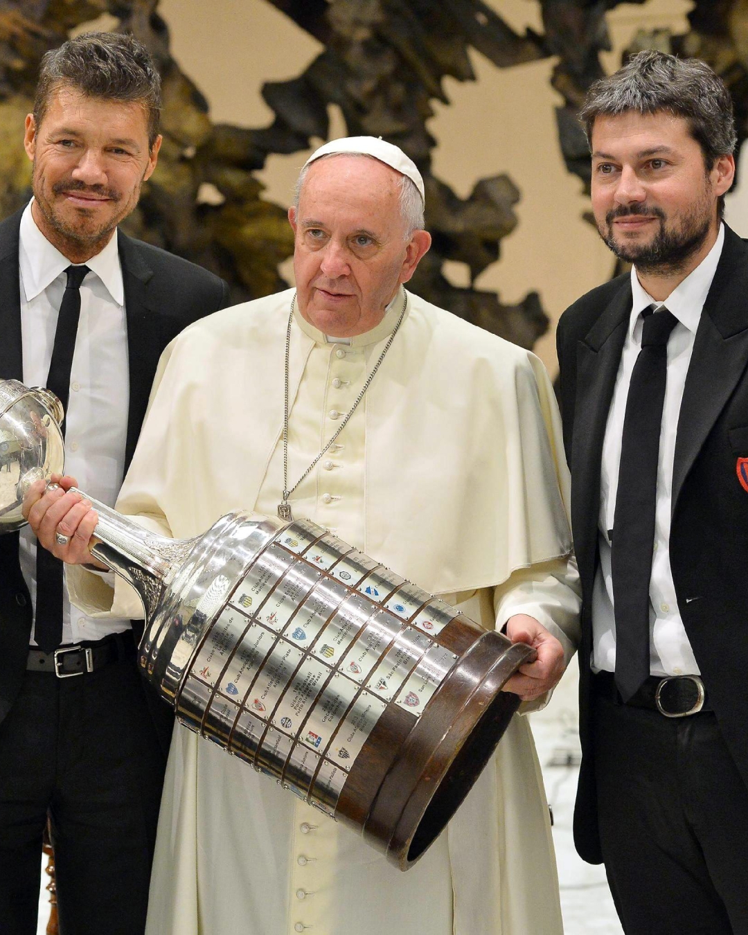 Tres hombres posan con un gran trofeo de fútbol. El hombre en el centro viste atuendo papal