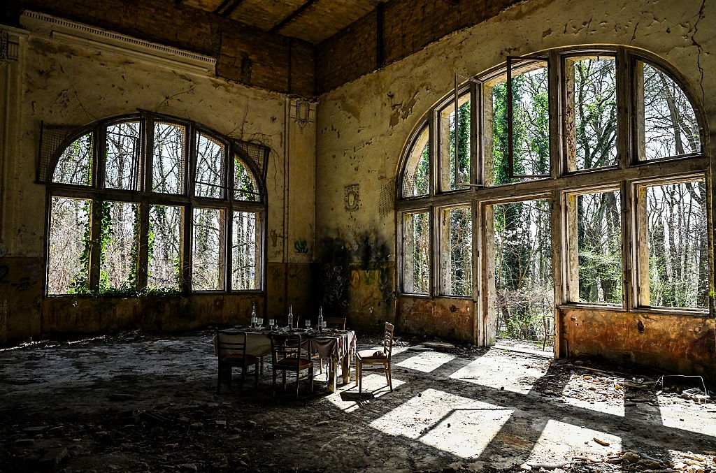 Abandoned grand room with large arched windows. Overgrown vines visible outside. A small, dusty dining table with chairs sits in the sunlight