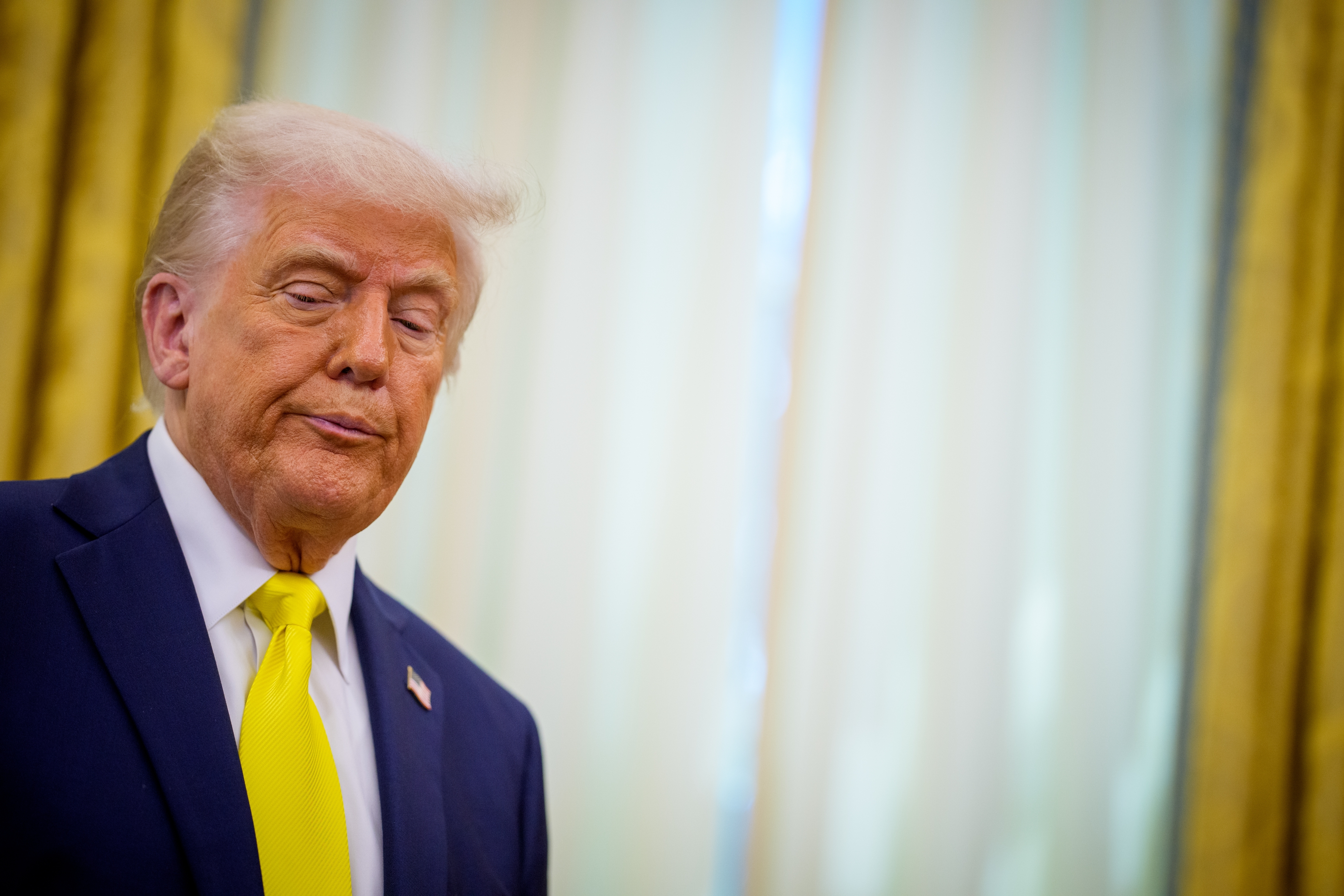 Person in a suit with a yellow tie stands indoors, looking down. Neutral backdrop with draped curtains