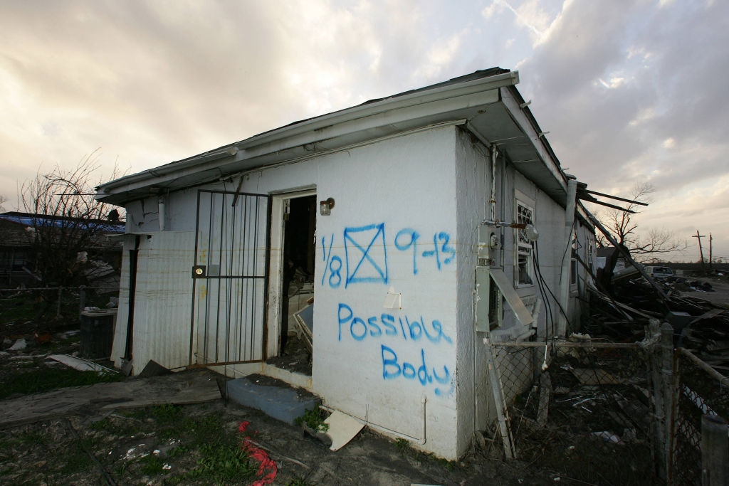 Abandoned building with graffiti marking stating "Possible Body" on exterior wall, surrounded by debris and a damaged entrance