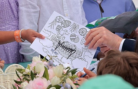 A group of children showing coloring pages to a man seated among them at a table decorated with flowers