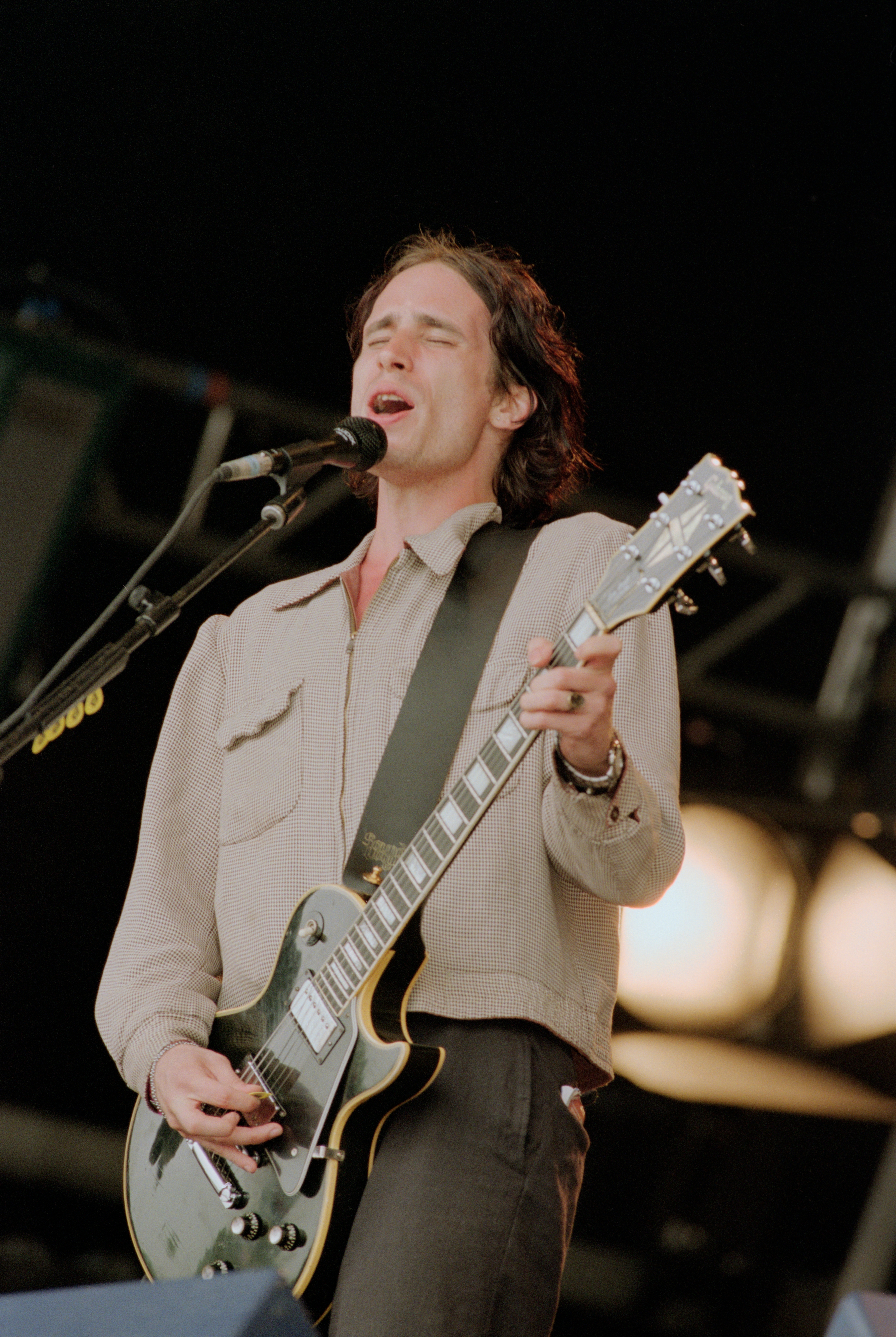 A person passionately sings and plays guitar on stage with eyes closed, wearing a casual button-up shirt