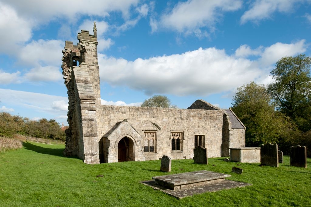 Ancient stone church ruins with a partially collapsed tower, surrounded by gravestones on a grassy field under a cloudy sky