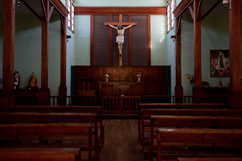 Interior of a small church with wooden pews, crucifix above the altar, and religious statues on either side