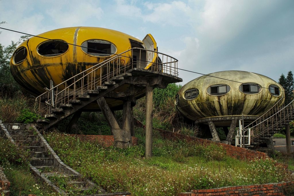 Two dirty and faded UFO-shaped houses on stilts with stairs, set in a grassy area. One door is open, suggesting abandonment