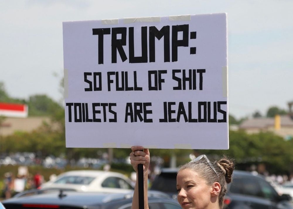 Person holding a protest sign that reads, &quot;TRUMP: SO FULL OF SHIT TOILETS ARE JEALOUS&quot; in a parking lot setting
