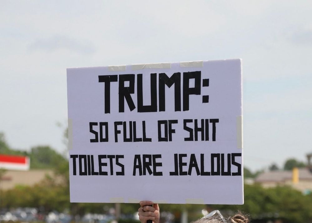 Person holding a protest sign that reads, "TRUMP: SO FULL OF SHIT TOILETS ARE JEALOUS" in a parking lot setting