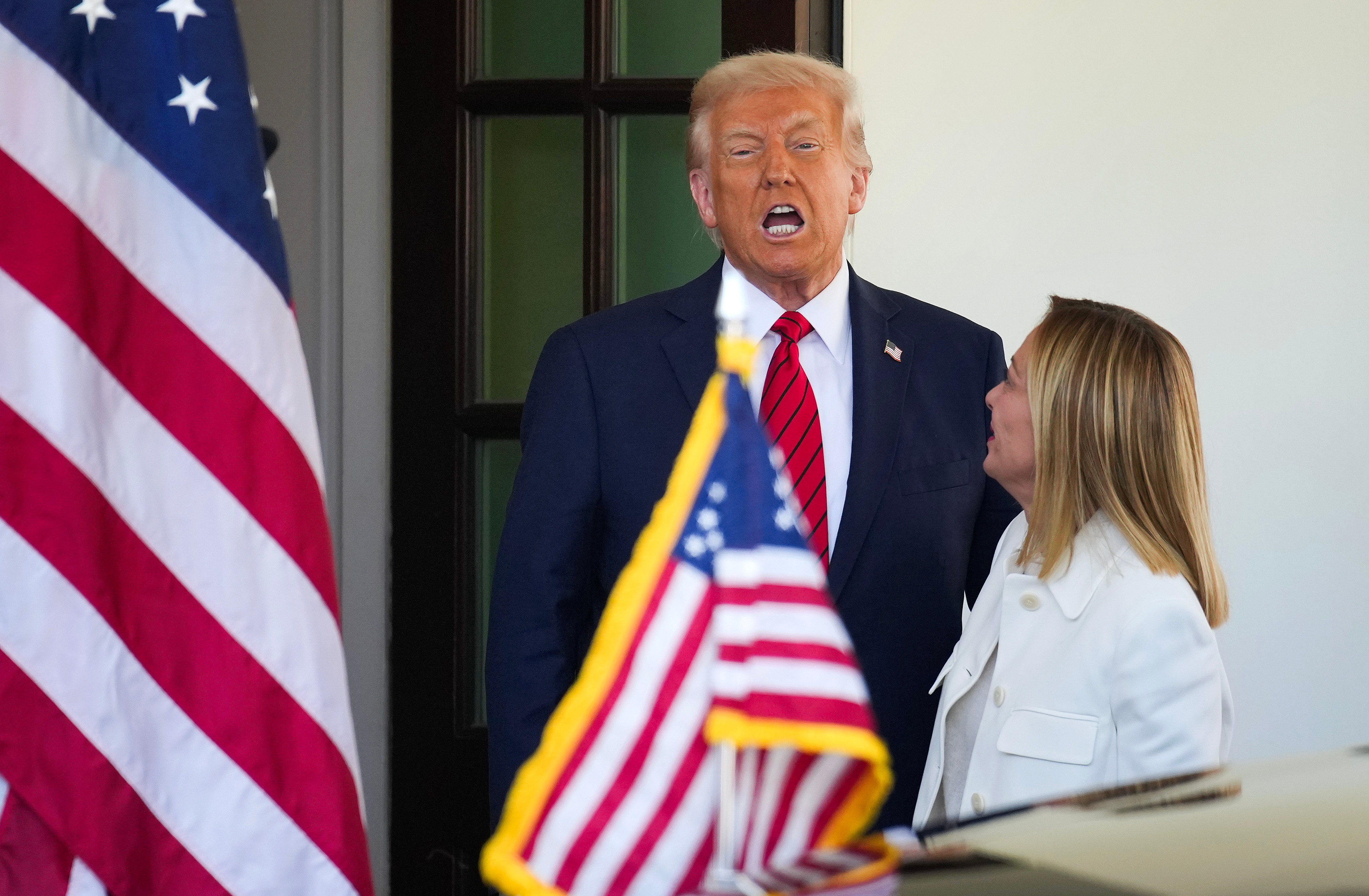 Two individuals, one in a suit with a striped tie, next to an American flag, outdoors at what appears to be an official or formal event