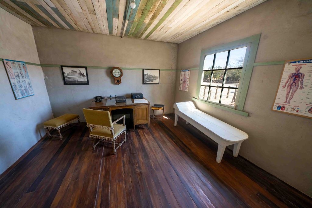 Vintage-style office with wooden floors, a desk with typewriter, two chairs, a bench, wall clock, and anatomical charts on the walls