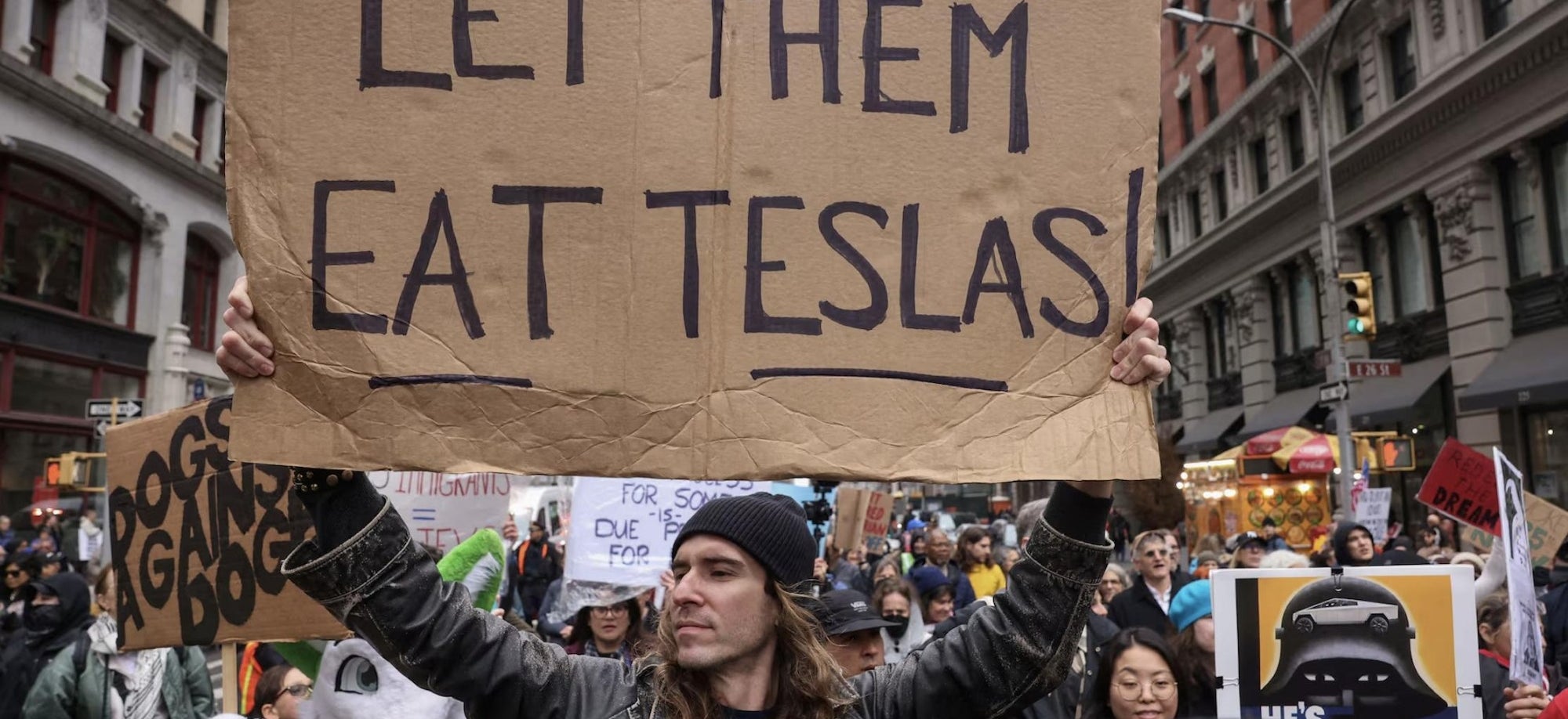 Person holding a cardboard sign reading &quot;LET THEM EAT TESLAS!&quot; at a crowded protest in an urban setting