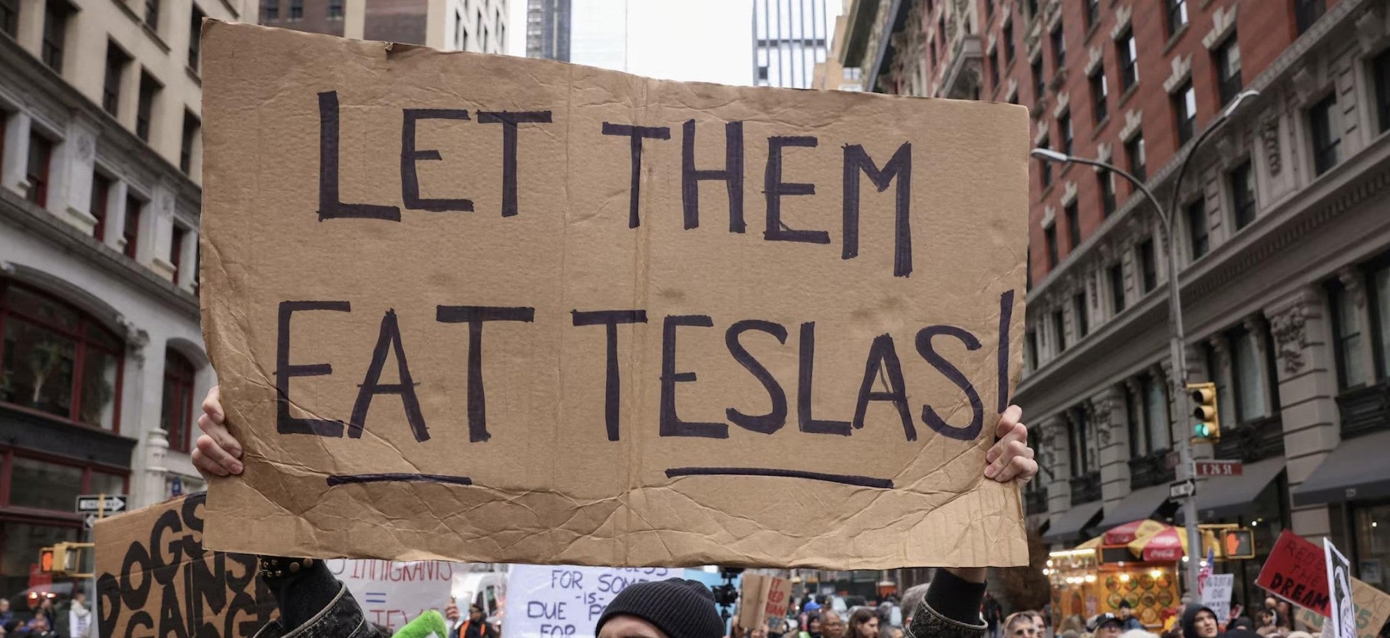 Person holding a cardboard sign reading "LET THEM EAT TESLAS!" at a crowded protest in an urban setting
