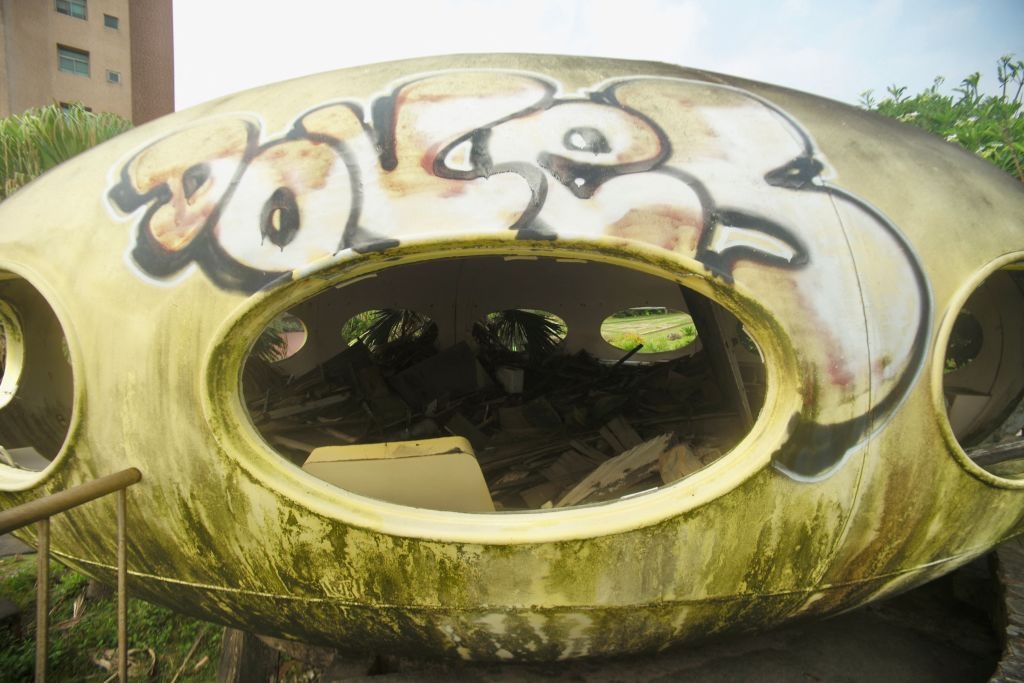 Graffiti-covered, egg-shaped structure with multiple oval windows, surrounded by greenery. Interior appears cluttered with debris