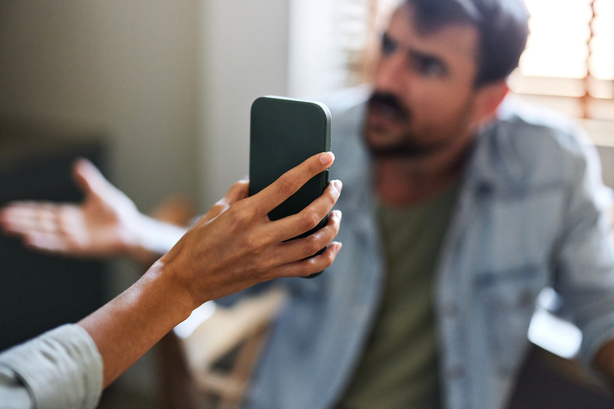 Person holding phone confronts another person gesturing in frustration. Focus on phone, possibly indicating a tense conversation about messages