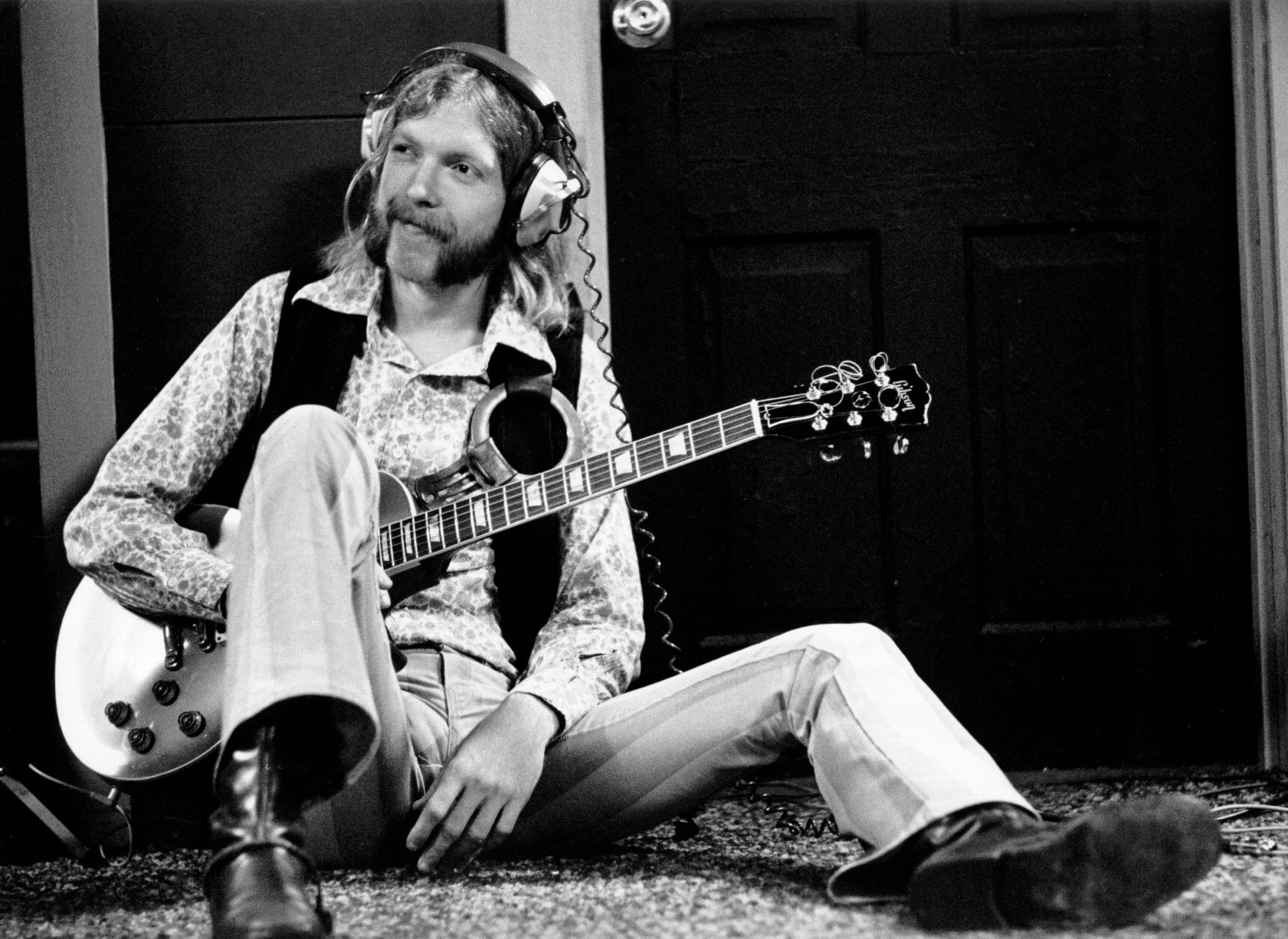 A man sits on the floor with headphones on, holding a guitar, appearing relaxed and casual in vintage attire