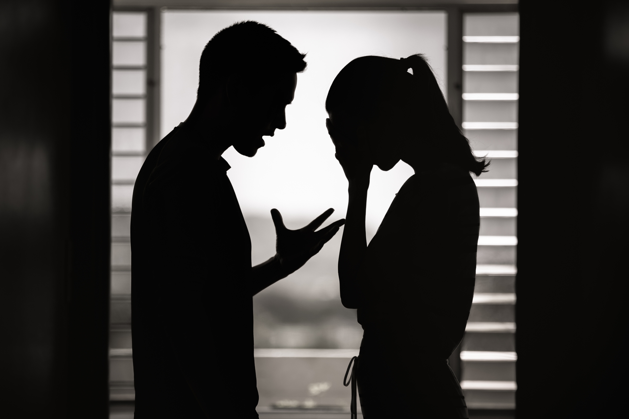 Silhouetted couple arguing, showing frustration and distress, in front of a window with slatted blinds