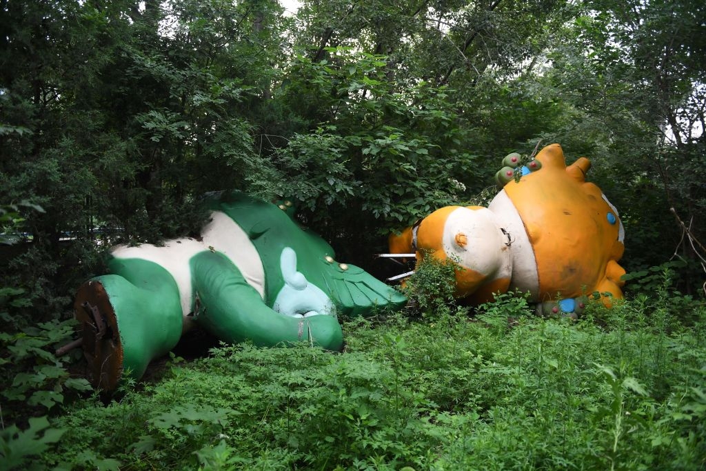 Large, overgrown statues of colorful, cartoon-like animals lie abandoned in dense greenery, partially hidden by foliage