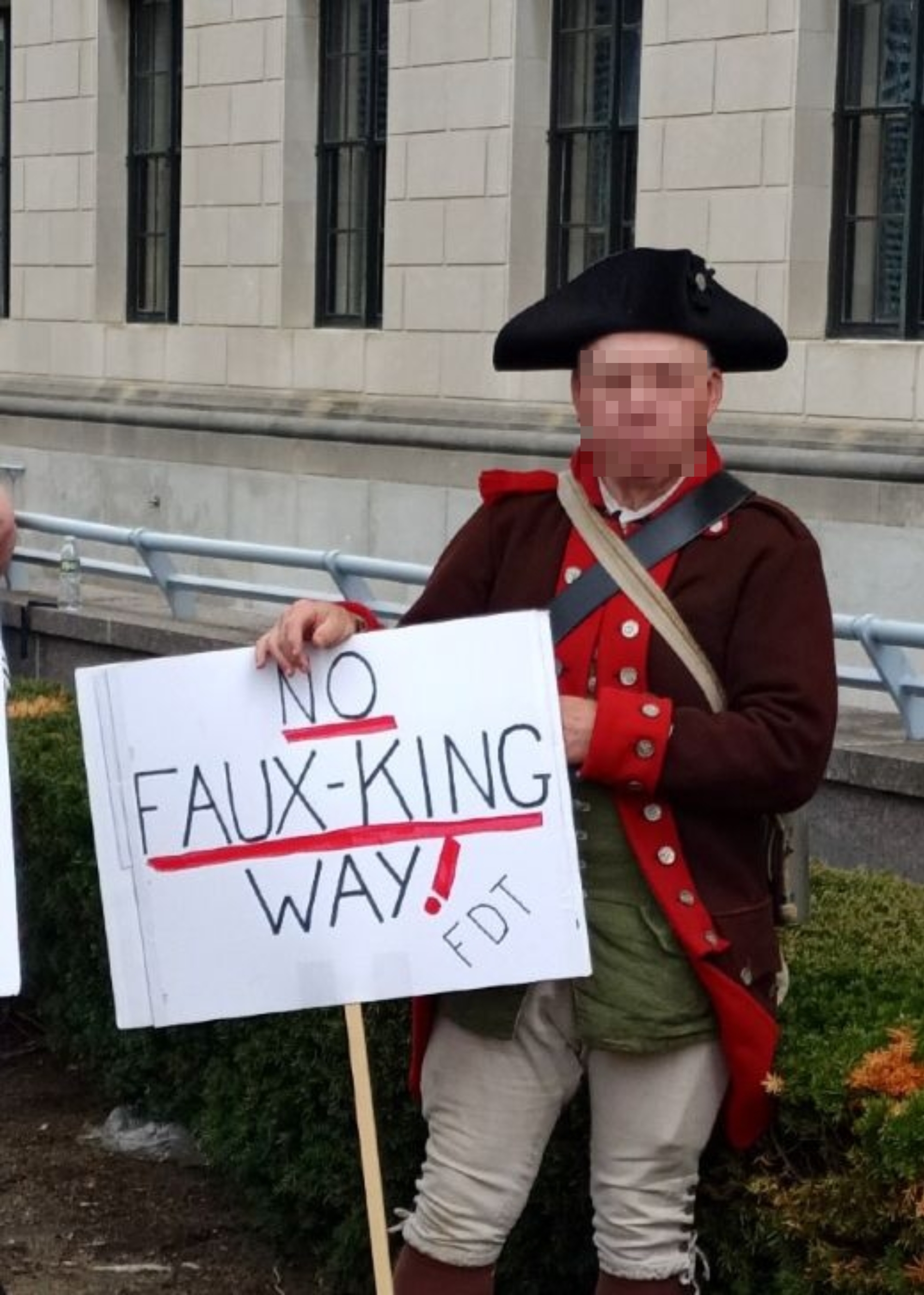 A man in colonial attire holds a protest sign reading "No Faux-king Way!"