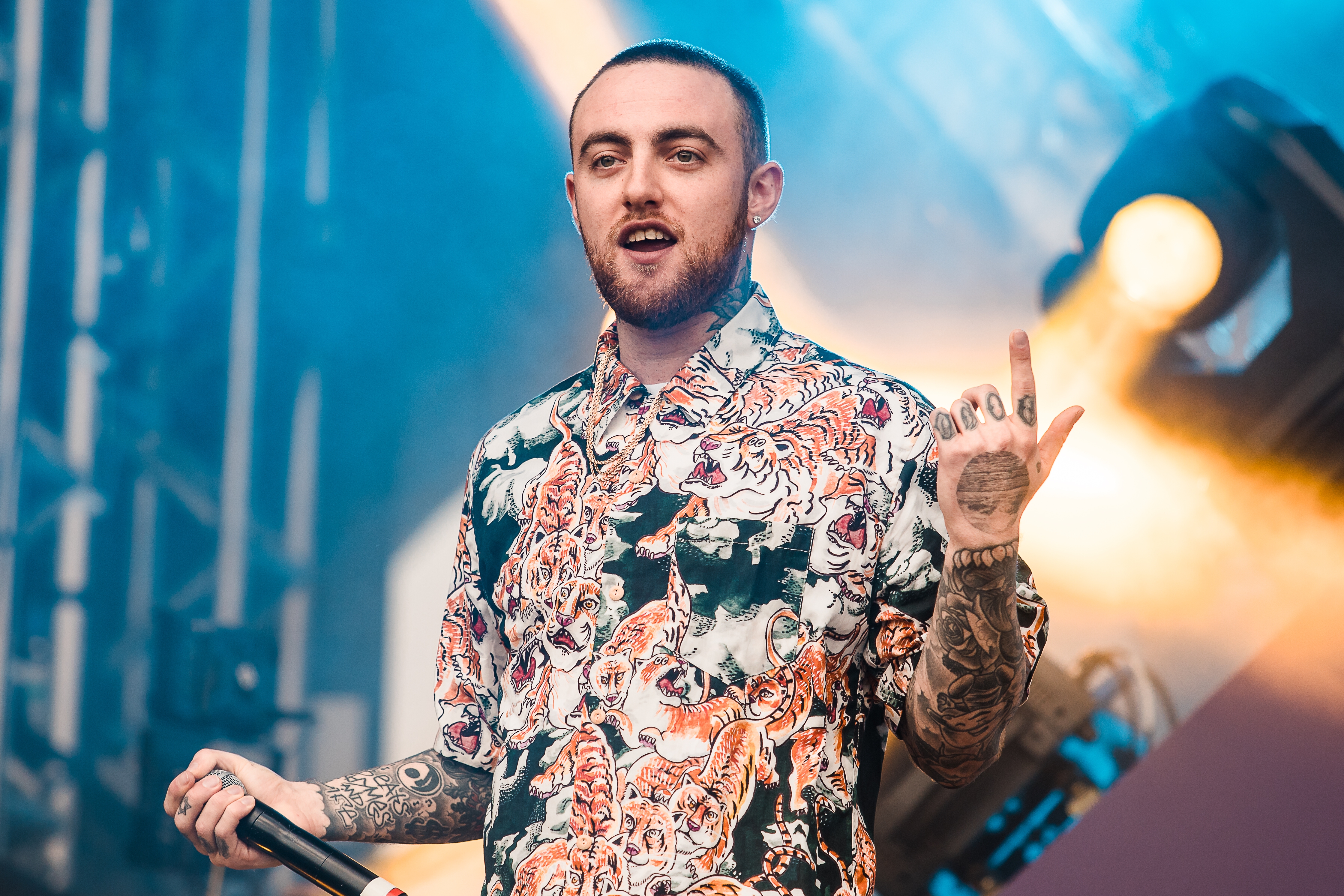 A person performs on stage, wearing a patterned shirt with fierce animal motifs, holding a microphone, with tattoos visible on their arms