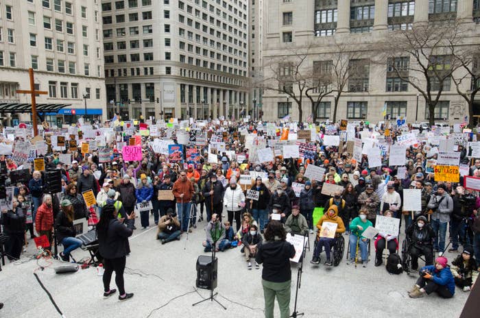 Large crowd at a city protest, participants hold signs demanding political and social change. Speakers address the audience from a small stage