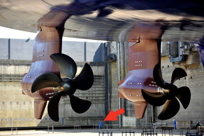 Massive ship propellers in dry dock, raised above the ground, with two workers standing beneath them for scale