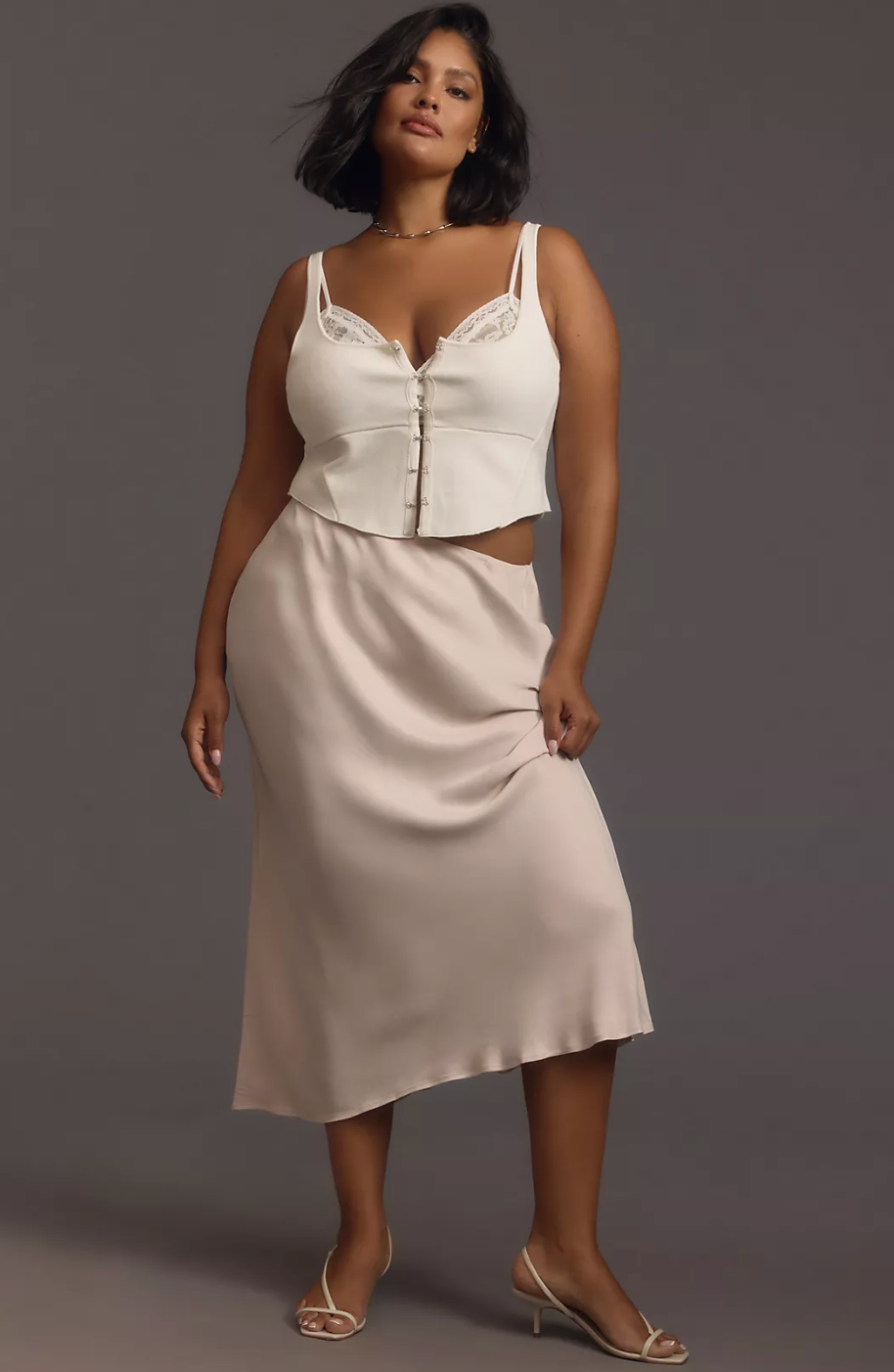 Model wearing a stylish sleeveless top and midi satin skirt, with strappy heels, posing confidently